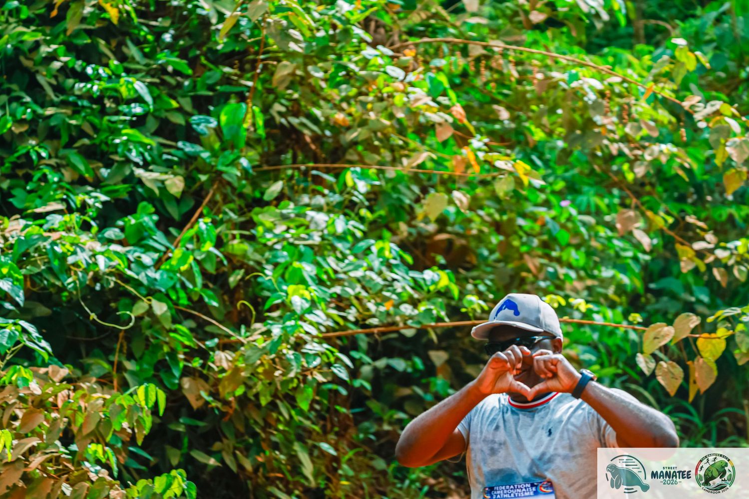A person in a hat makes a heart shape with their hands in front of lush green foliage.