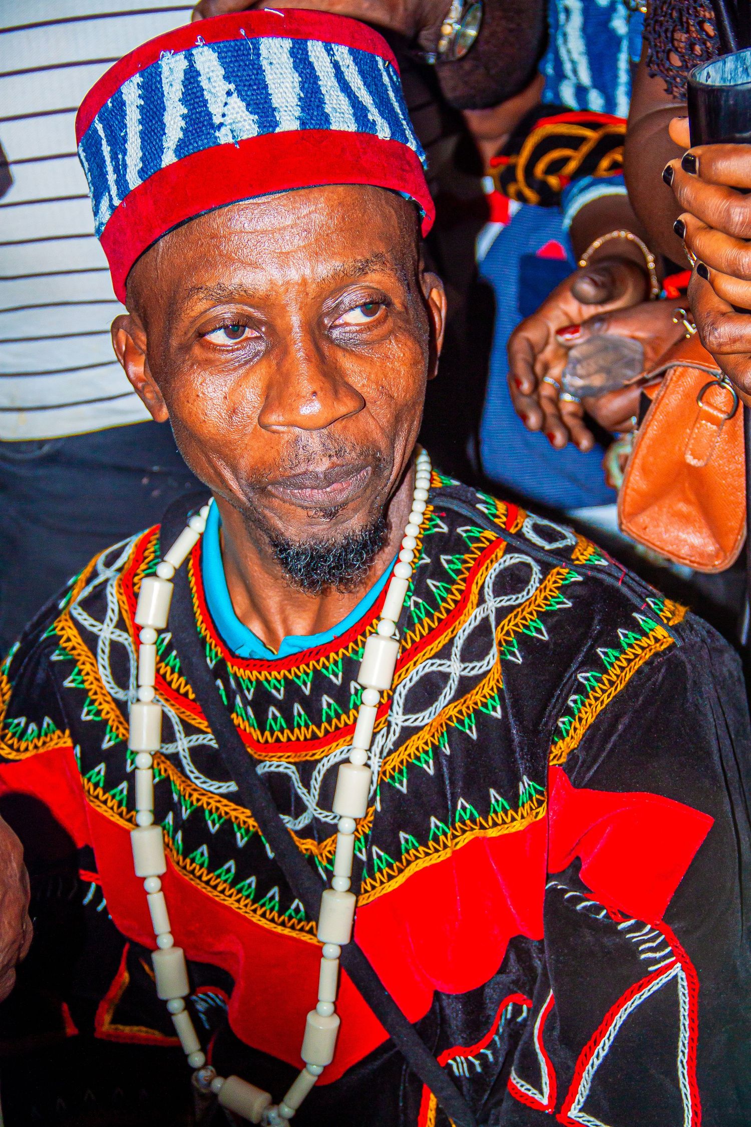 Elder African man in colorful traditional Cameroon attire with beaded necklace and blue patterned cap.