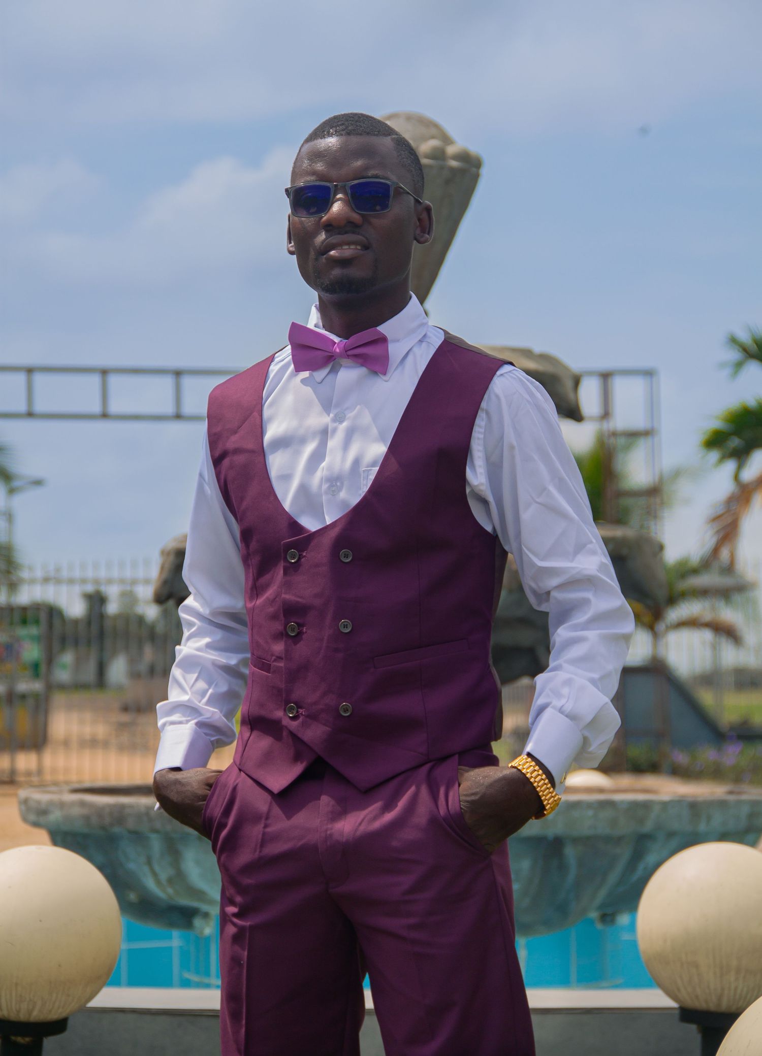 Stylish man in purple vest suit with bow tie and sunglasses posing outdoors.