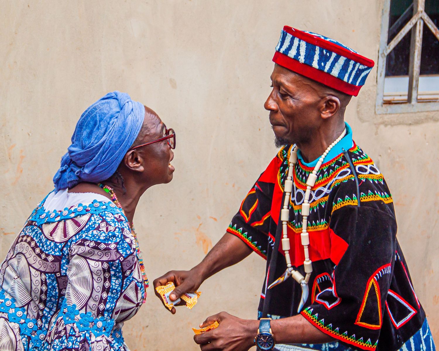 Two African adults in traditional attire having a conversation outside a building.