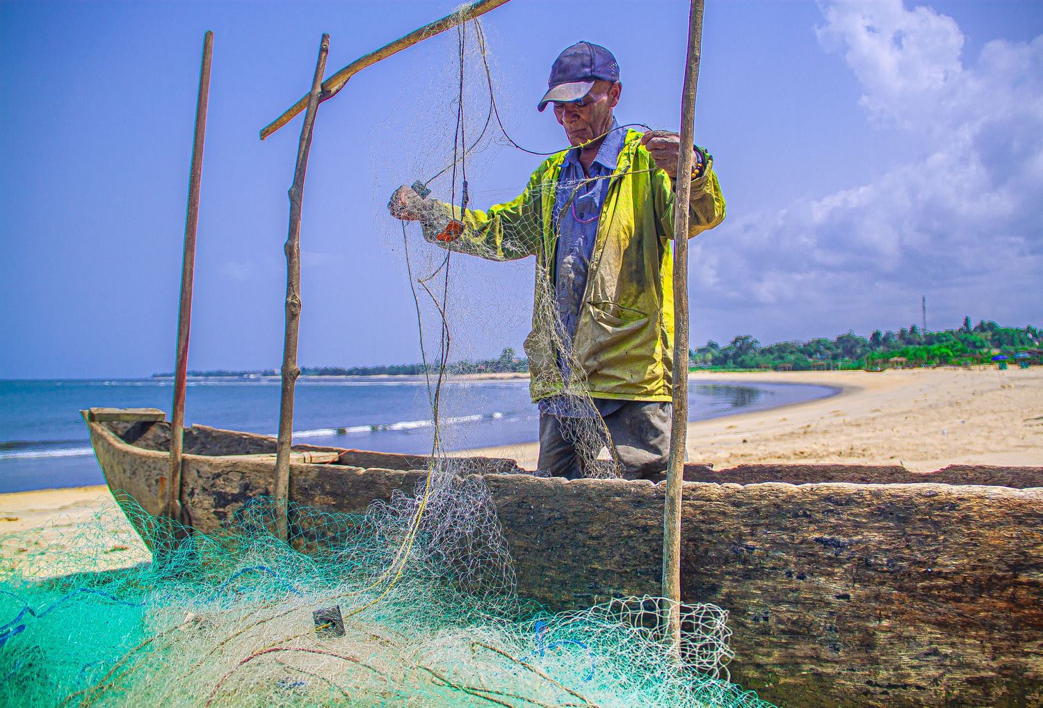 Fisherman in green jacket handling turquoise fishing nets beside a wooden boat on a tropical sandy beach.