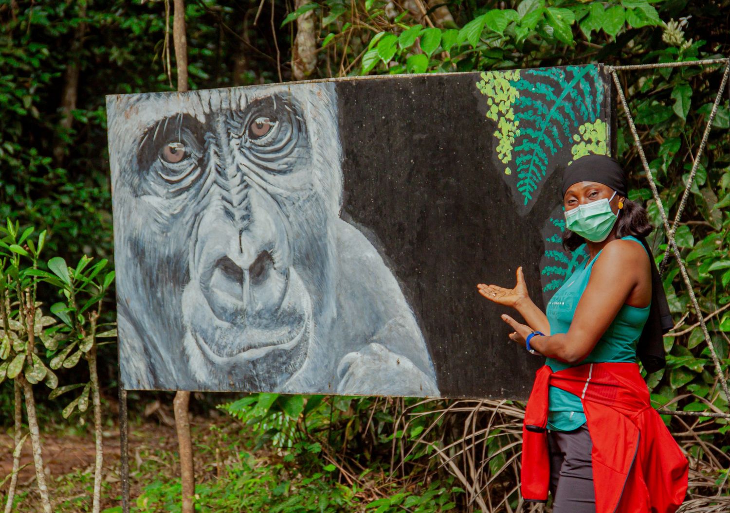 Woman in face mask standing beside a painted gorilla portrait artwork in a tropical forest setting.