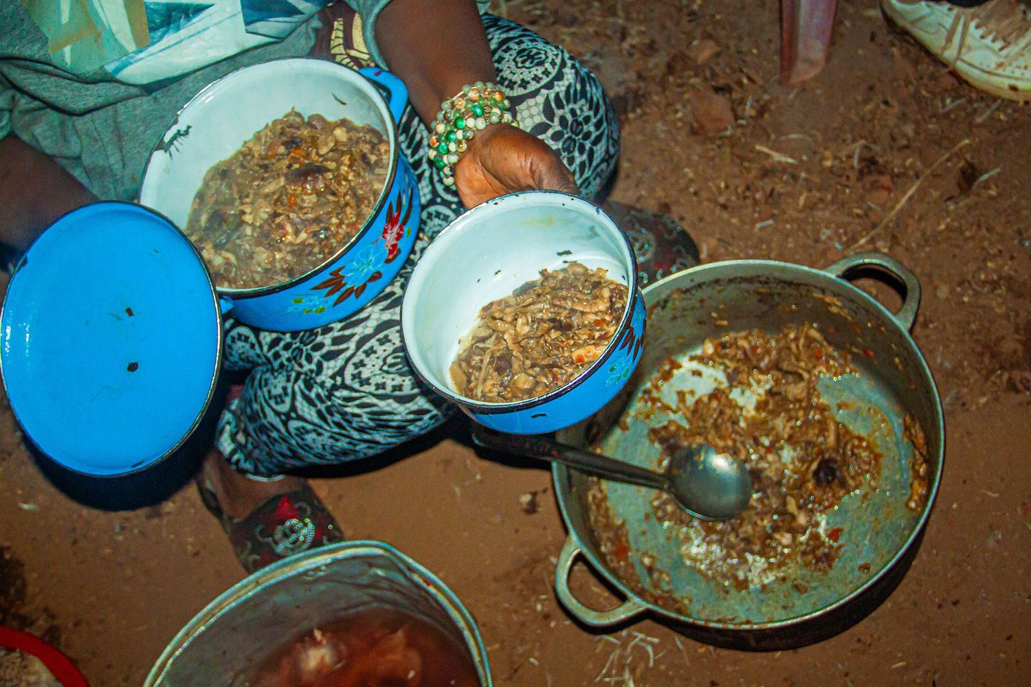 Bowls of traditional African food with meat and grains served in blue and metal pots on red dirt ground.
