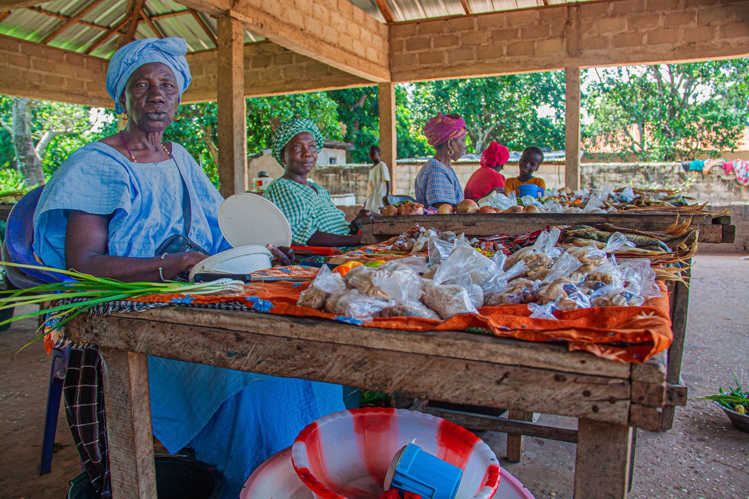 African market women in traditional attire selling food and goods at a wooden stall.