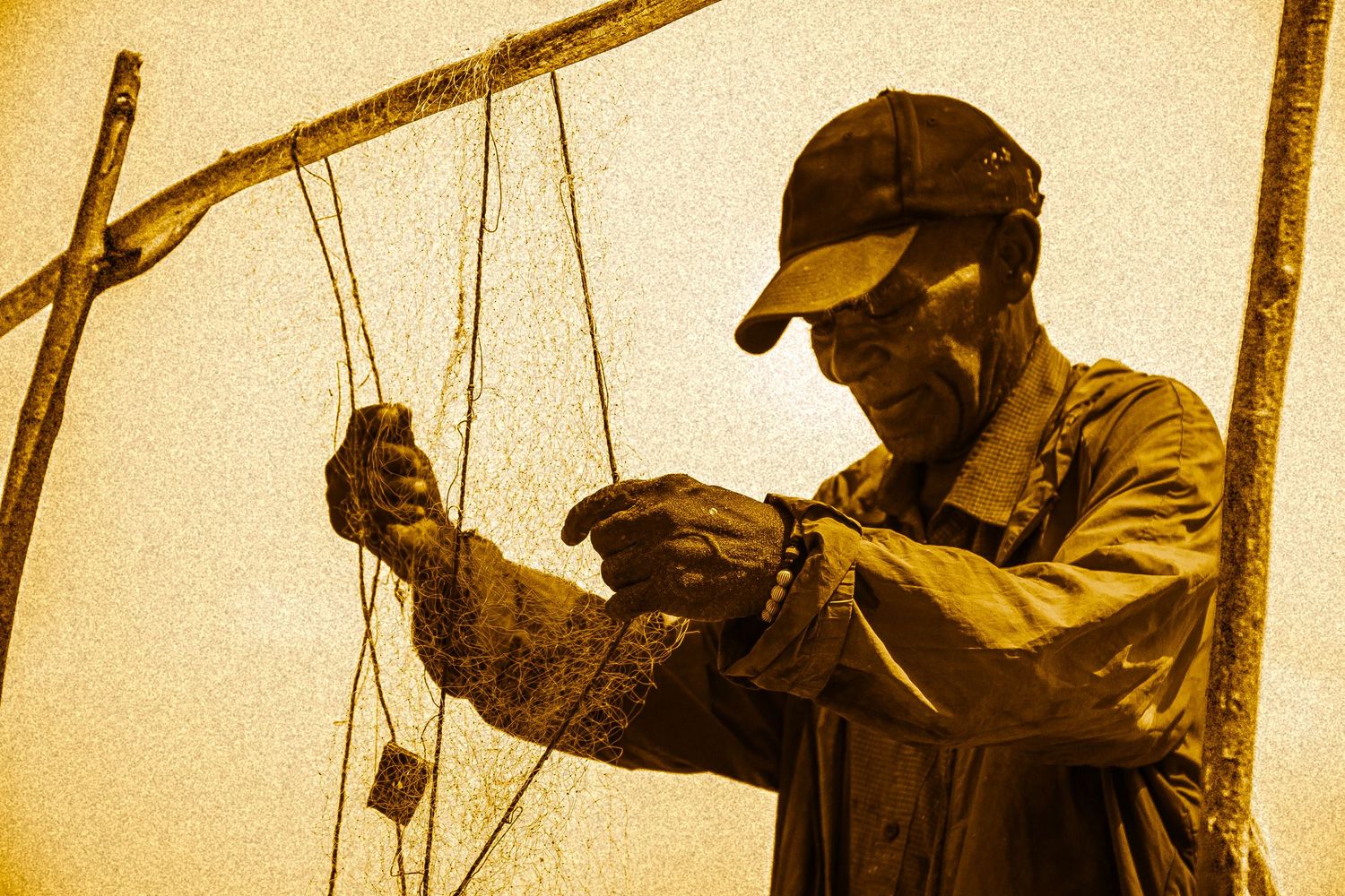 Weathered fisherman in cap carefully untangles fishing nets outdoors in sepia-toned photograph.
