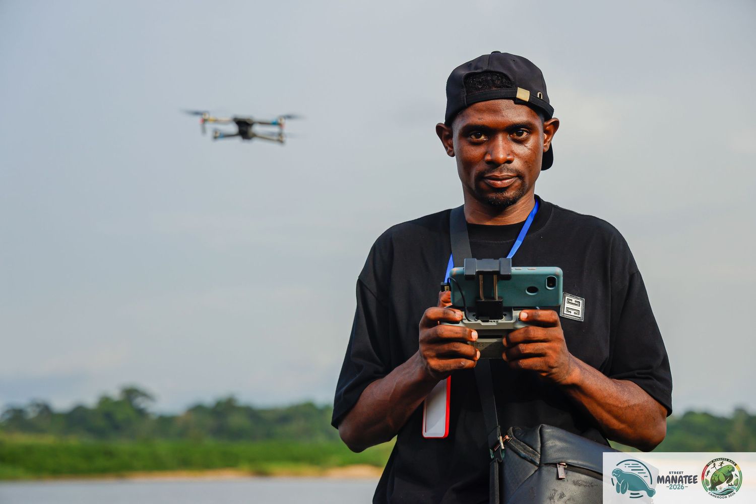 A man operates a drone controller at a riverside location in Africa under cloudy skies.