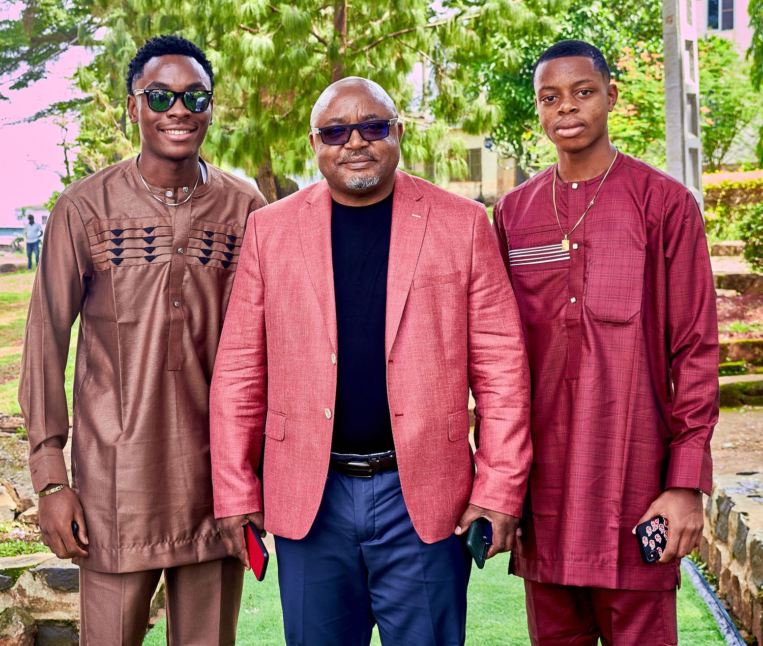 Three men dressed in elegant traditional African attire pose together outdoors for a formal portrait.