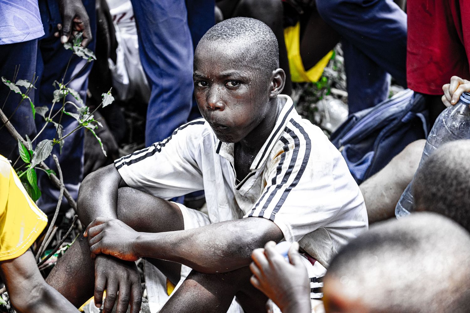 Young African boy sitting outdoors with knees up, looking thoughtful in a crowd.
