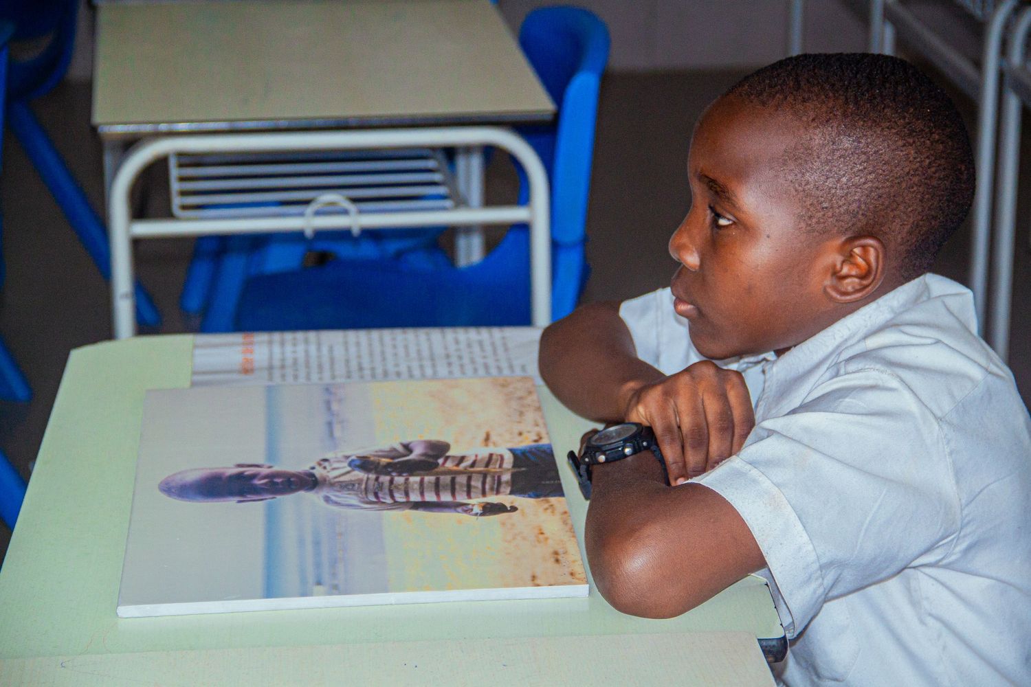 A young African child reads a book at a school desk in a bright classroom.