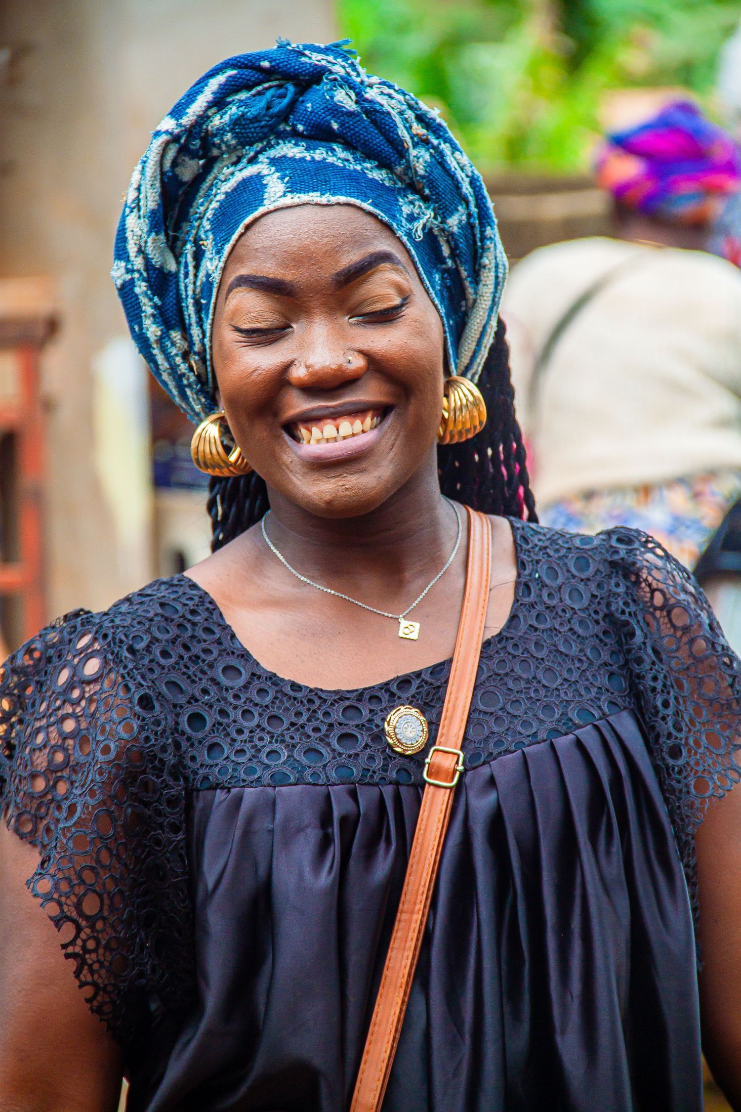 A smiling African woman in a blue head wrap and black lace top wearing gold earrings outdoors.