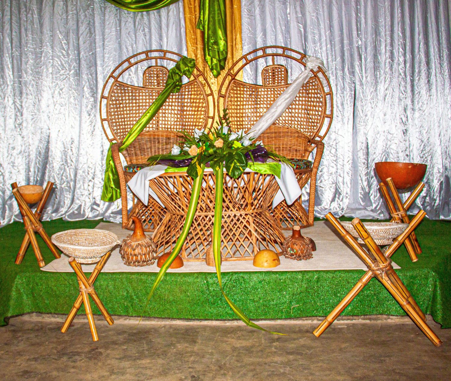 Elegant wicker peacock chairs decorated with tropical flowers on a green-draped stage for a wedding ceremony.