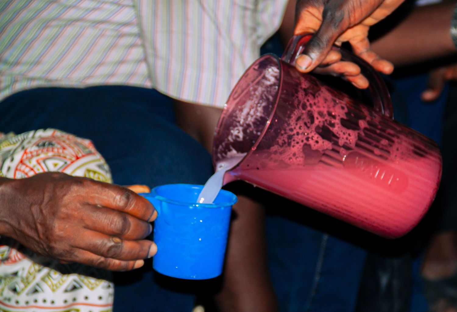 Person pouring a pink beverage from a large pitcher into a small blue cup at a gathering.