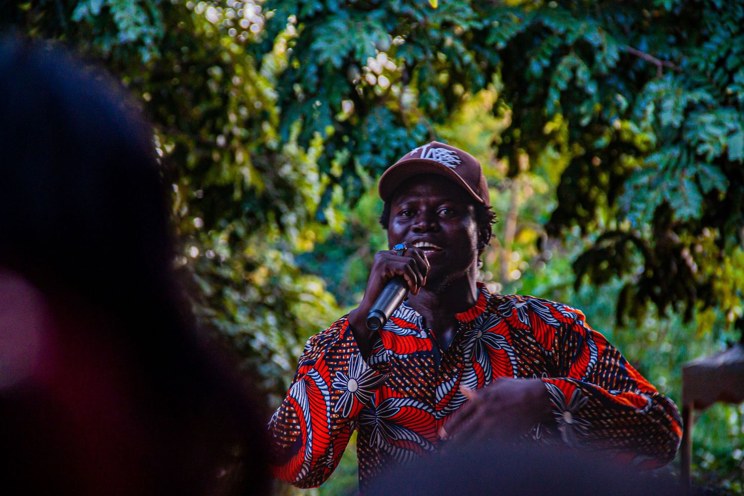 African man in vibrant red patterned shirt speaking into microphone outdoors among trees.