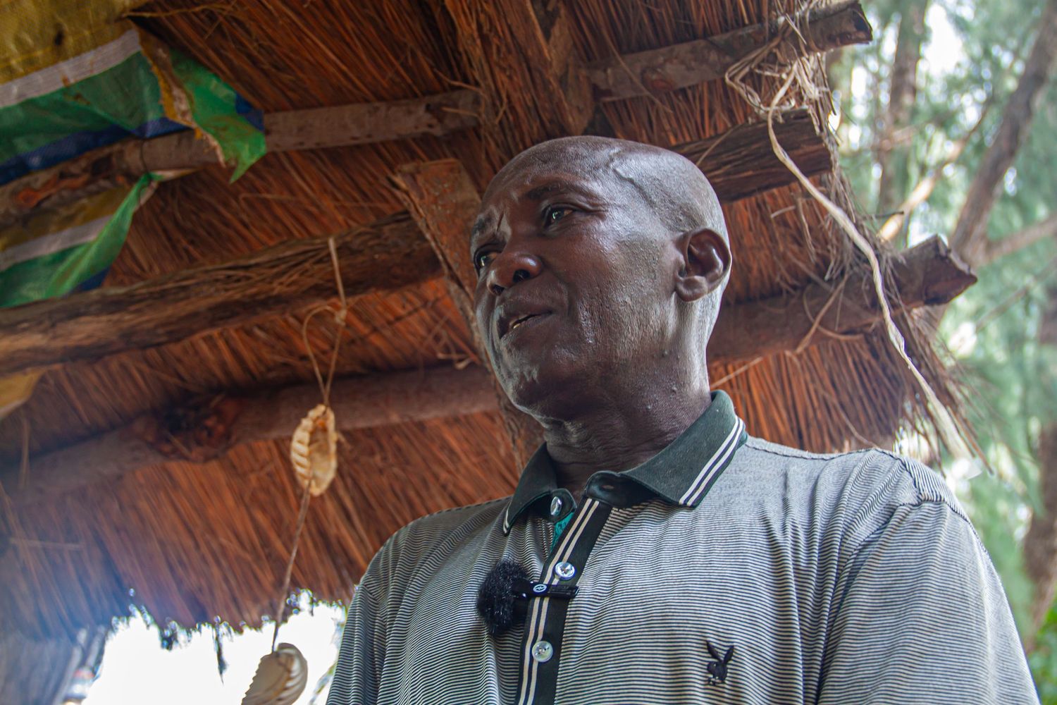 A middle-aged African man stands thoughtfully under a thatched roof with hanging decorative objects behind him.