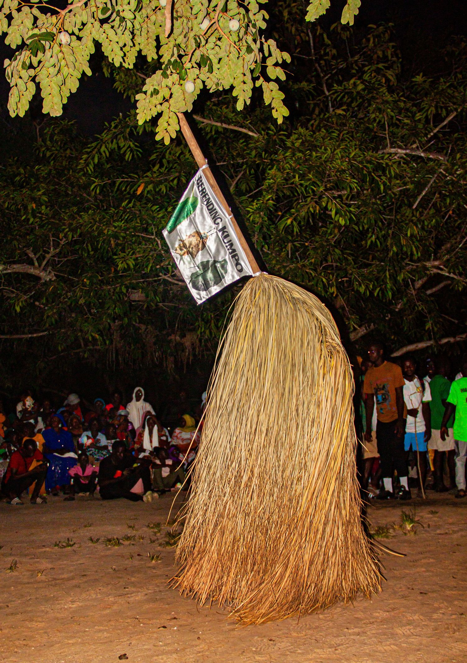 A traditional African straw costume performer holds a flag during a nighttime cultural ceremony outdoors.