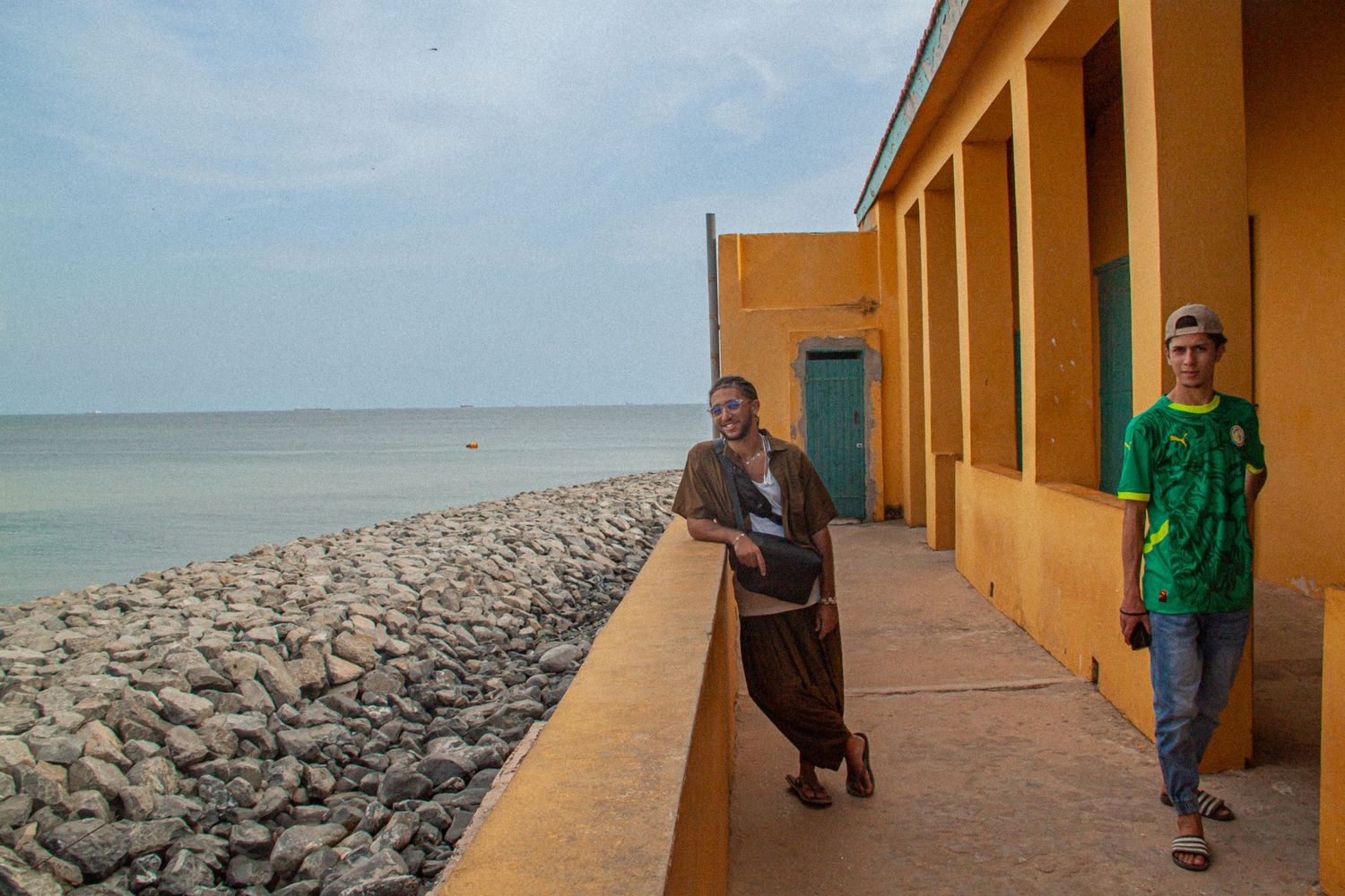 Two people standing on a seaside promenade next to yellow colonial buildings with rocky shoreline in West Africa.