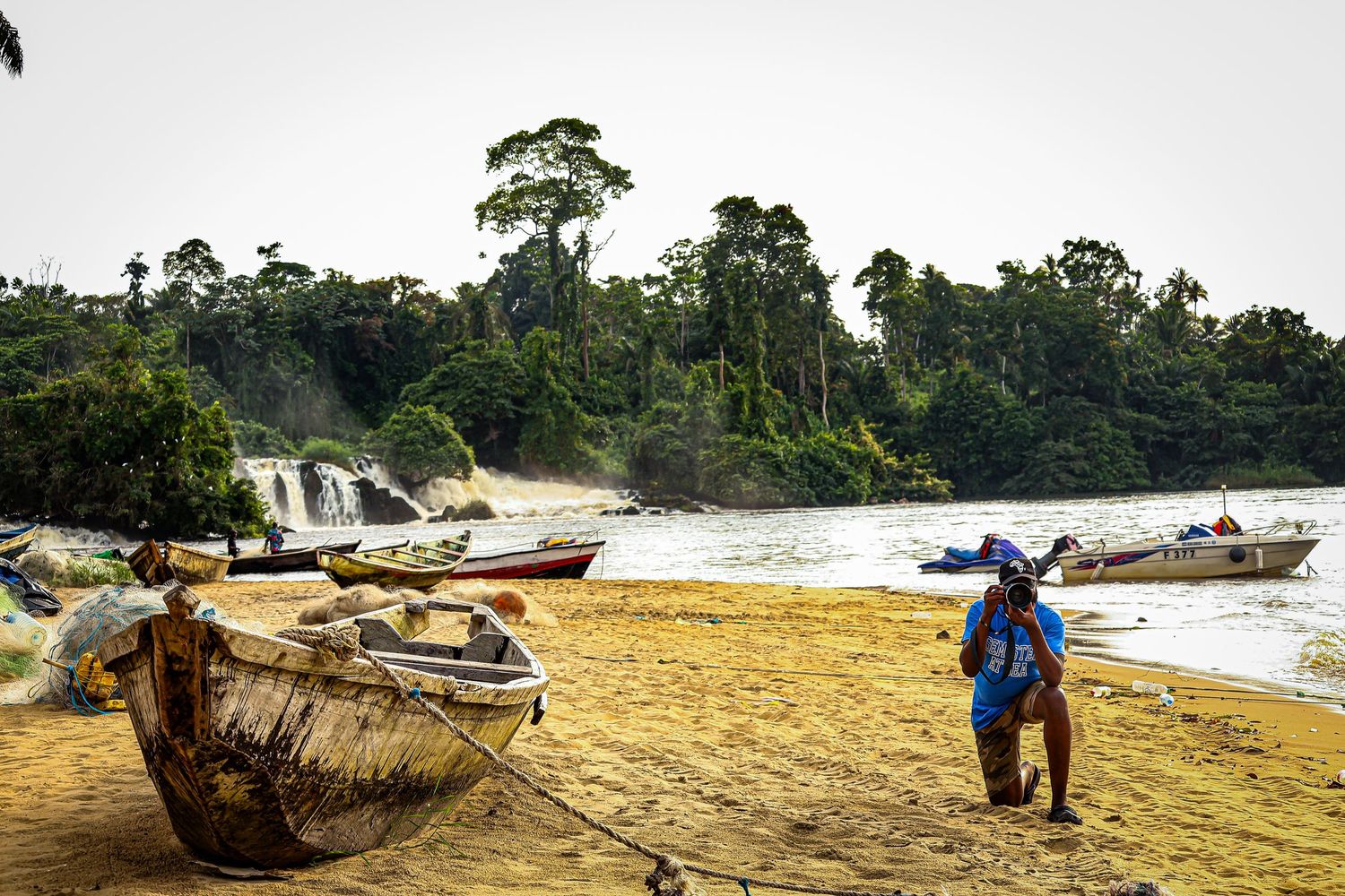 A fisherman stands on a sandy riverbank with wooden boats and a lush waterfall in the background in Africa.