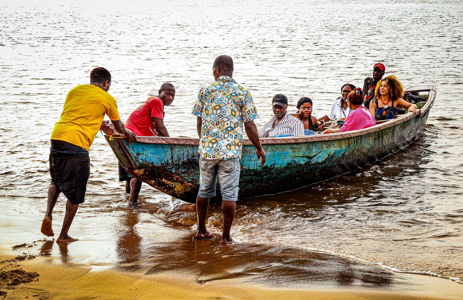 A group of people board a colorful wooden boat on a sandy shoreline in a tropical setting.