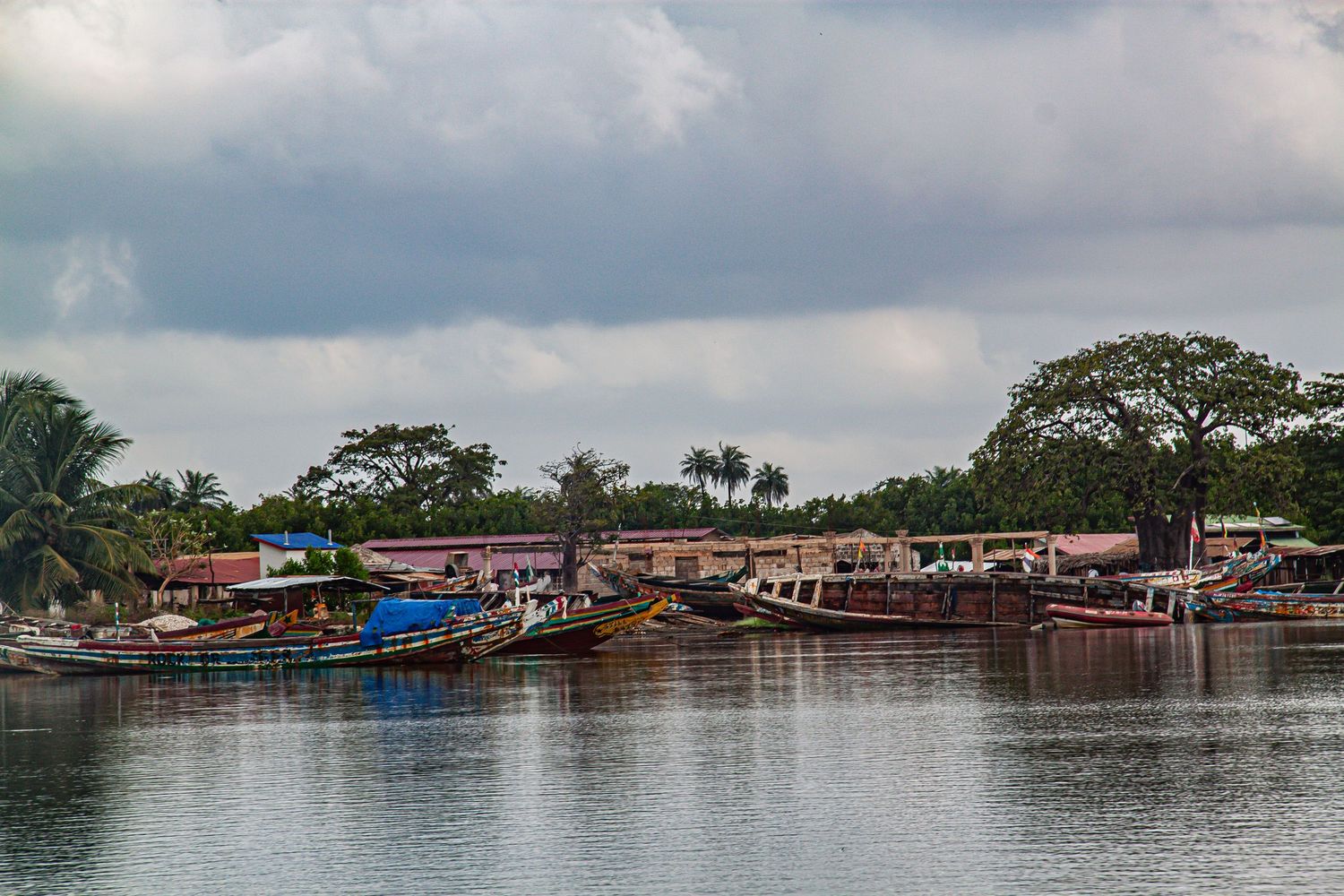 Colorful fishing boats docked at a calm river port surrounded by lush tropical trees under cloudy skies.