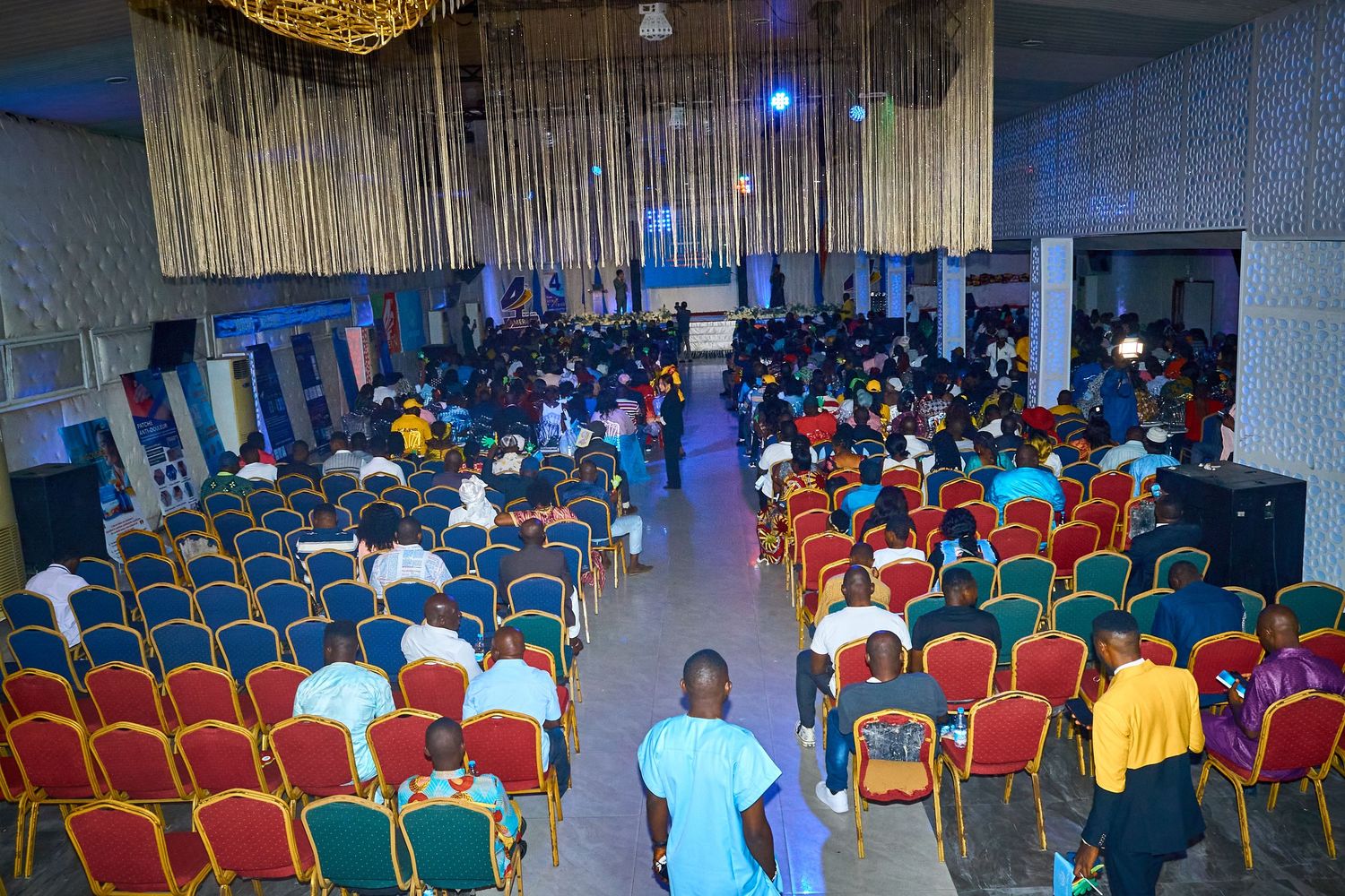 Large indoor event hall filled with rows of red and gold chairs and attendees.