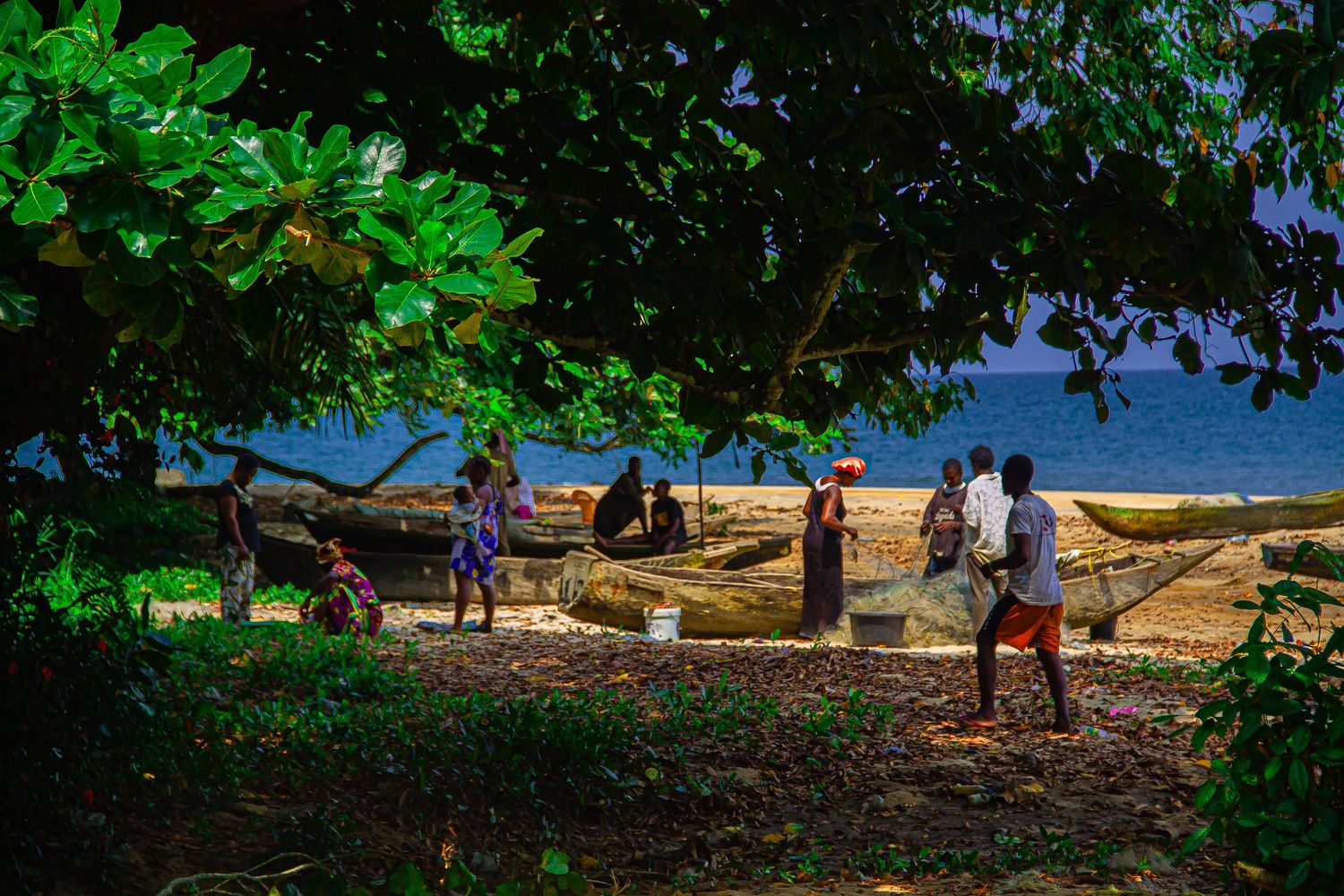 Local people gathered under large trees near wooden boats on a tropical beach shoreline.