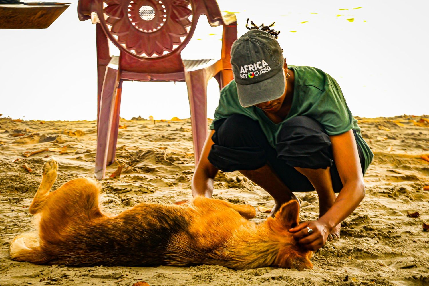 Man in green Africa shirt petting a brown dog rolling on sandy beach.