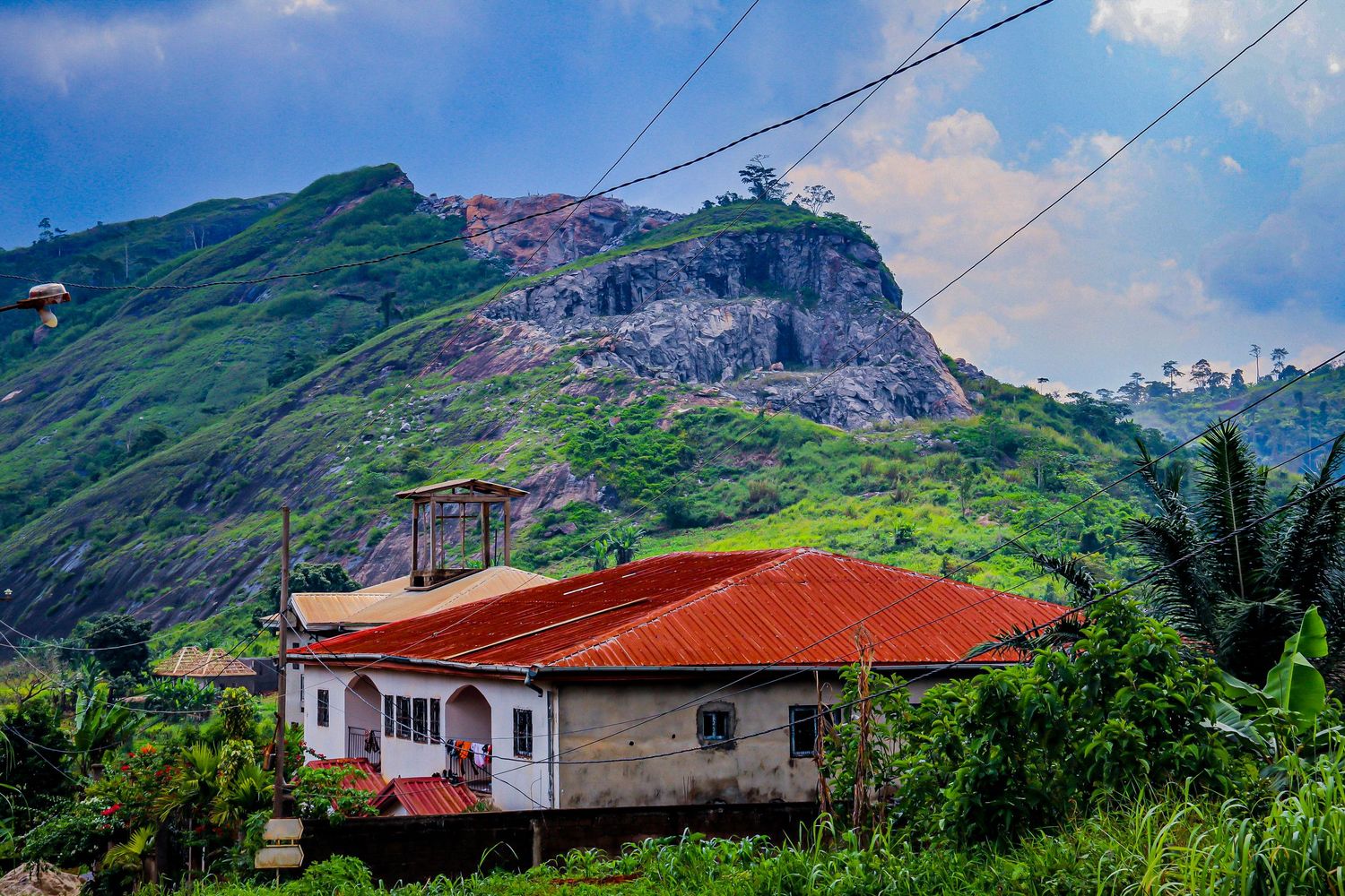 White house with red tin roof beneath a dramatic rocky mountain cliff in a tropical landscape.