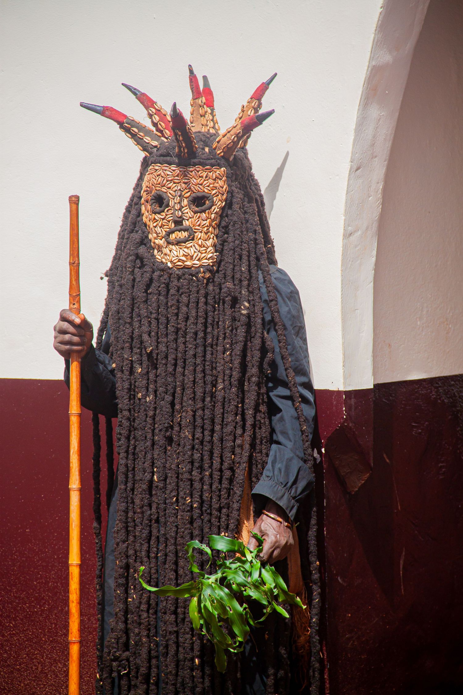 Person in traditional African ceremonial costume with woven mask, long dreadlock-like costume, holding staff and plants.