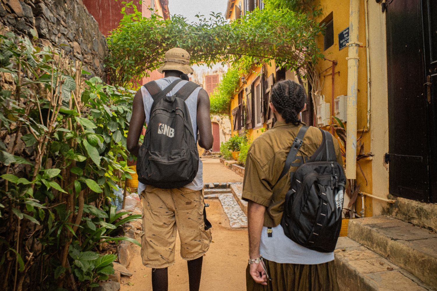 Two backpack-wearing tourists exploring a narrow colorful alley on Goree Island, Senegal.