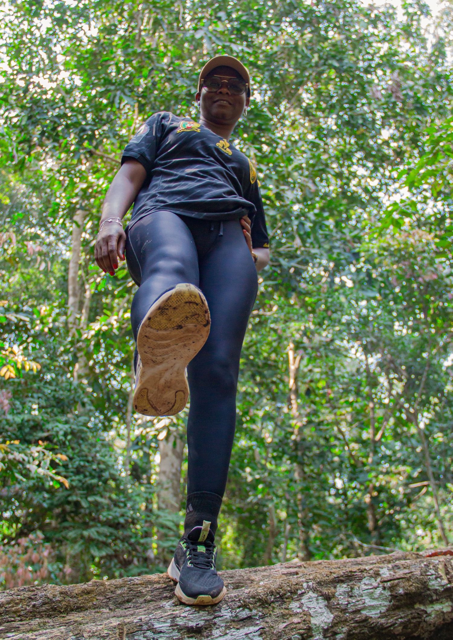 A person in black leggings and sneakers poses on a log in a lush green forest, kicking one leg forward.