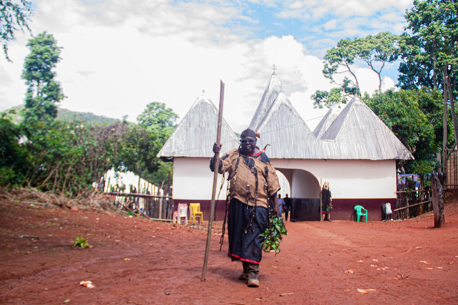 African man in traditional attire carrying a staff walks past white thatched-roof huts on red soil.