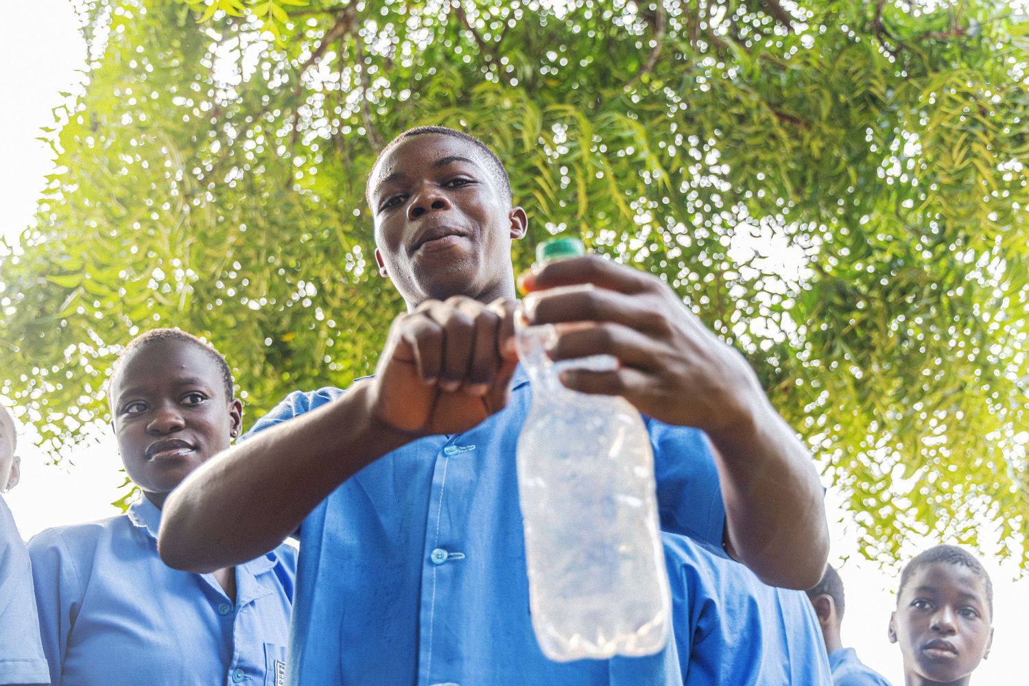 African students in blue uniforms examining a plastic bottle outdoors under trees.