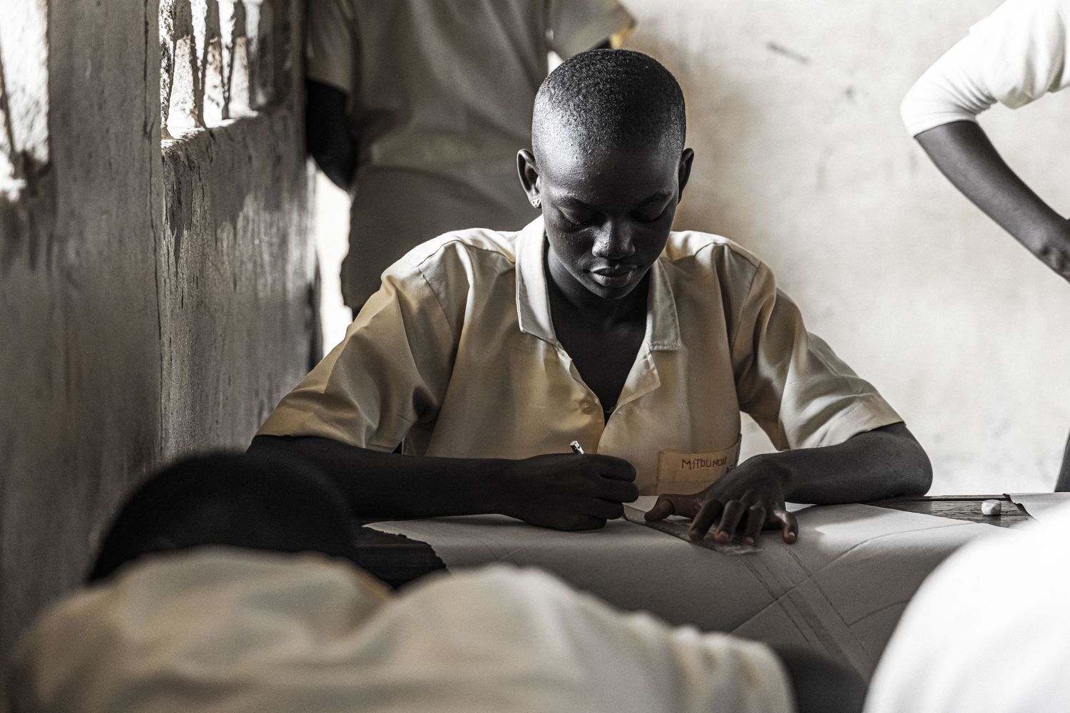 A young African boy focuses while writing or sewing at a workbench indoors.