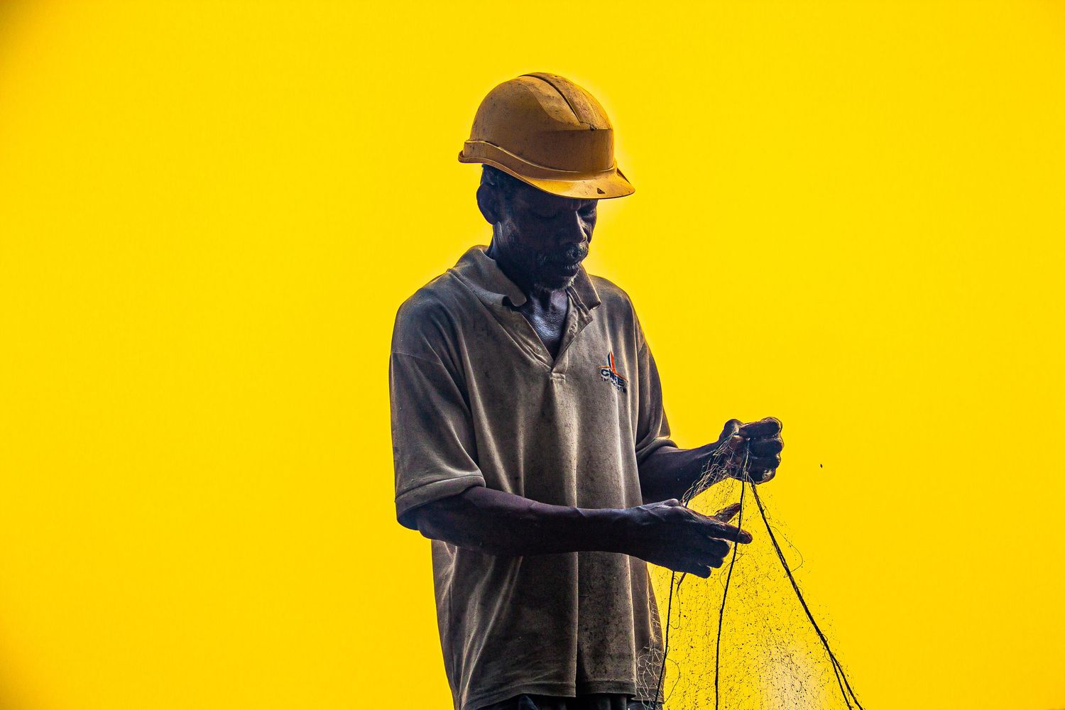 Silhouetted African worker in yellow hard hat holding fishing net against bright yellow background.