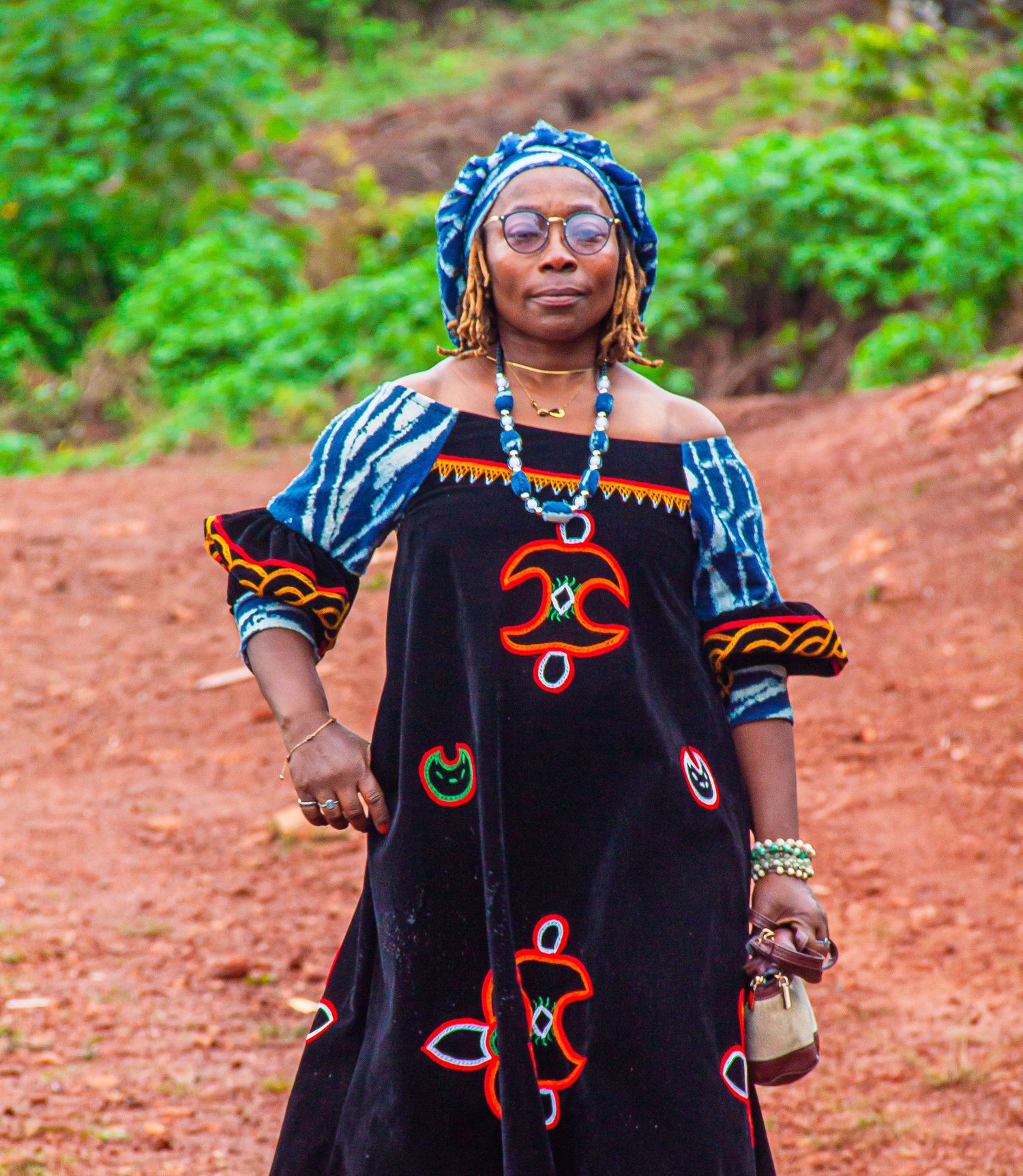 African woman in traditional black embroidered dress and blue head wrap walks on red dirt road.