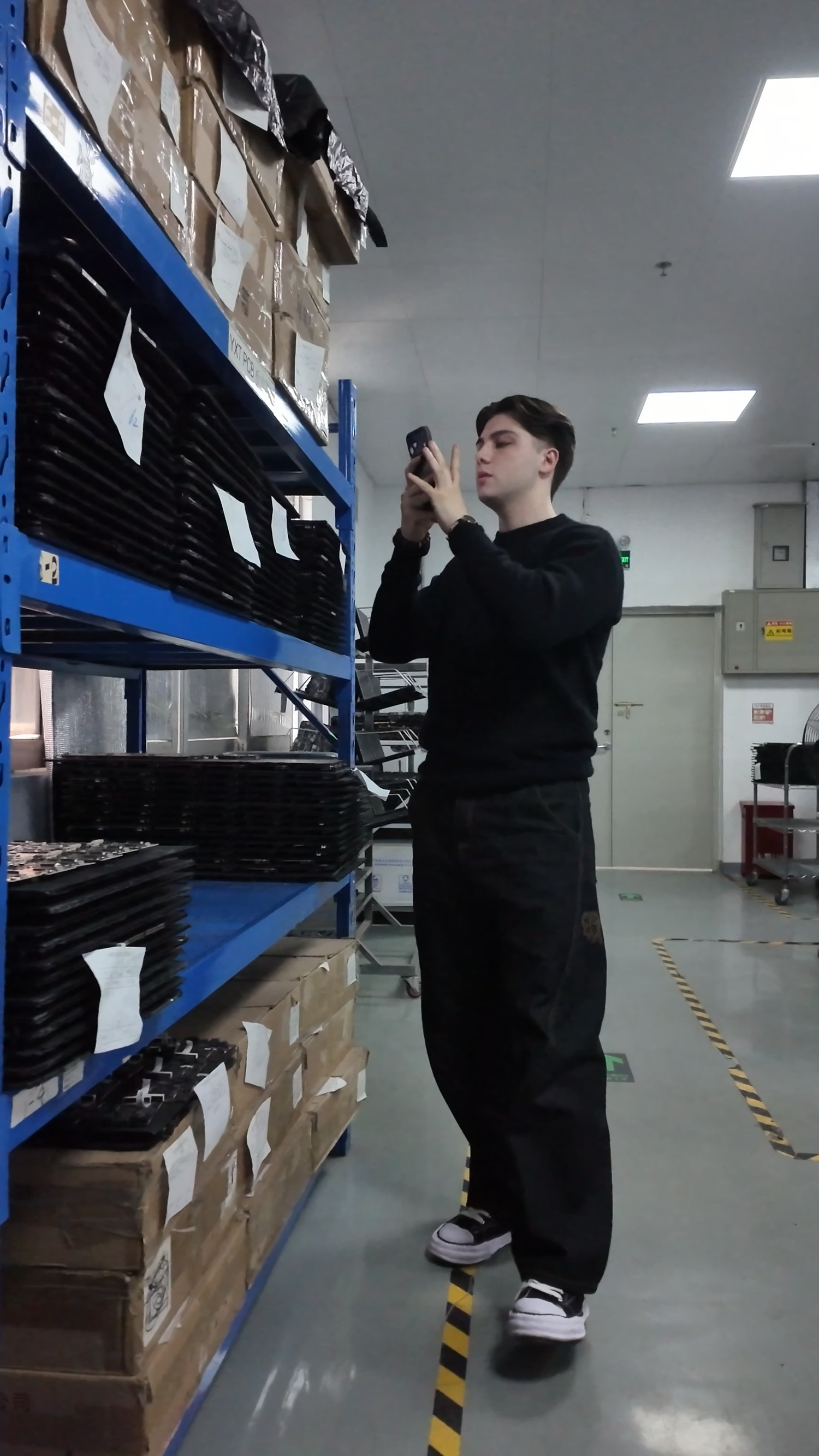 Young man photographing inventory on blue metal shelving racks in a modern warehouse facility.