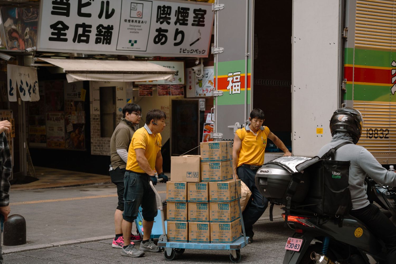 Workers in yellow uniforms unloading cardboard boxes on a cart in a Japanese street.