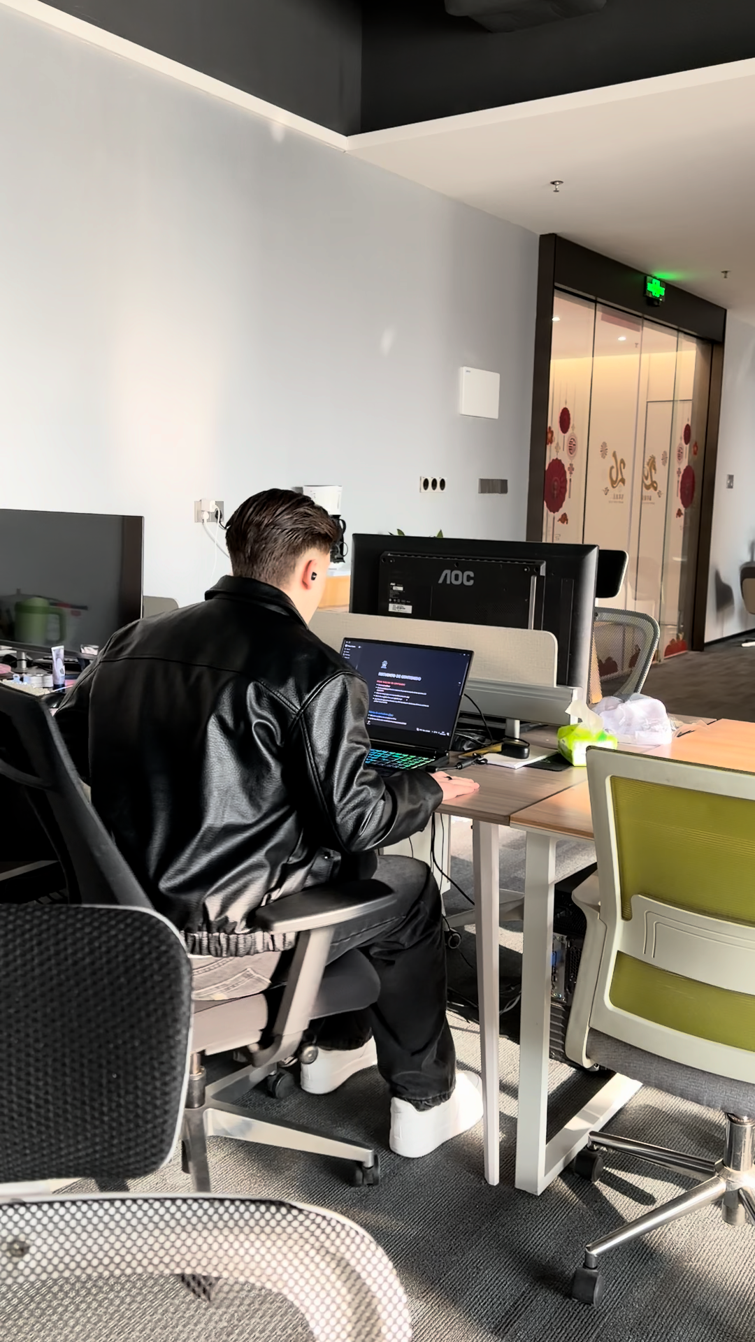 Person in black leather jacket working on a laptop at a standing desk in a modern office space.
