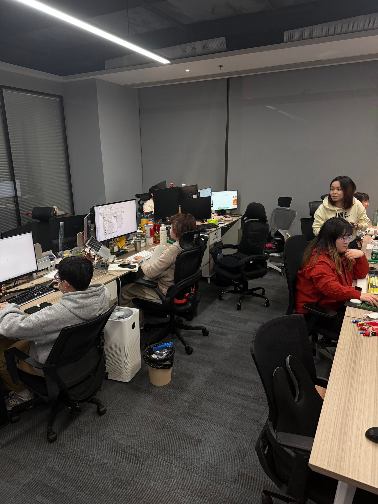 People working at computer desks in a modern office space with gray walls and bright overhead lighting.