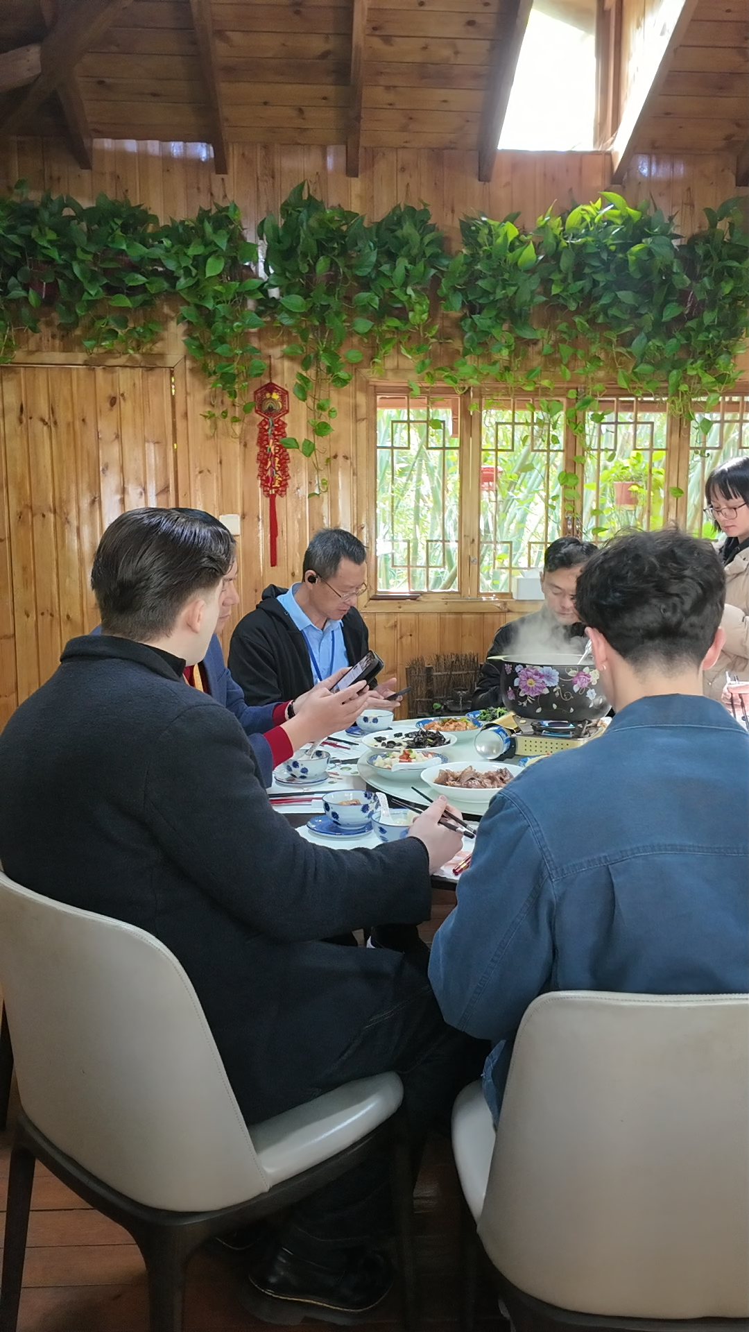 Group of people dining at a round table in a cozy wooden Chinese restaurant decorated with green vines.