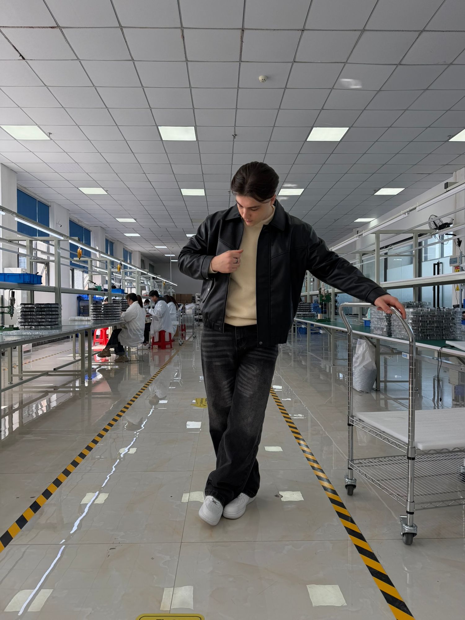 Young man in black jacket walking on factory floor with yellow safety markings.