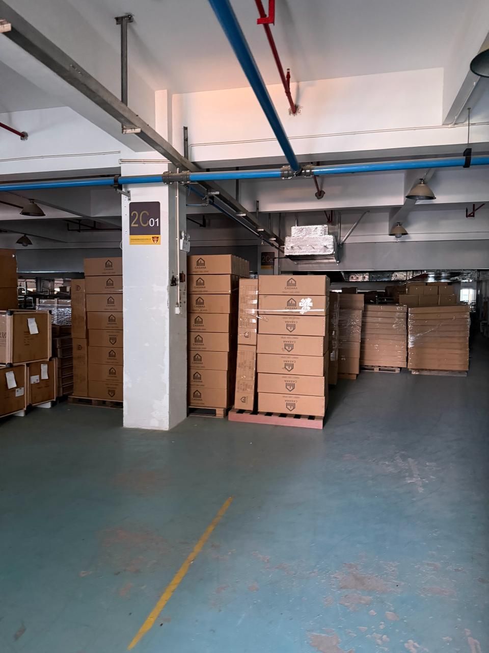Industrial warehouse interior with stacked cardboard boxes on pallets, blue pipes overhead, and a clean teal floor.