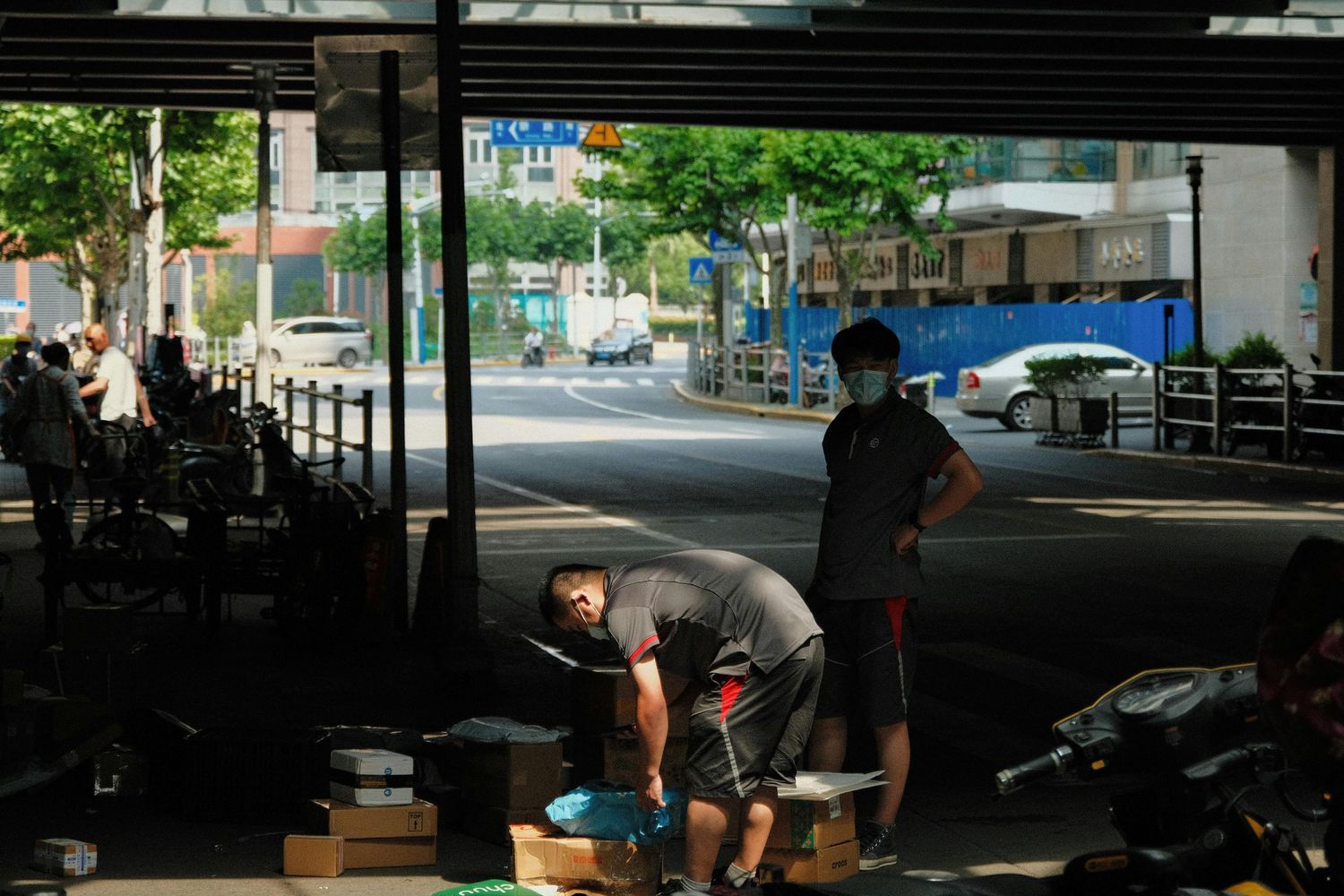 Two workers sorting and handling cardboard packages under a shaded urban overpass on a sunny day.