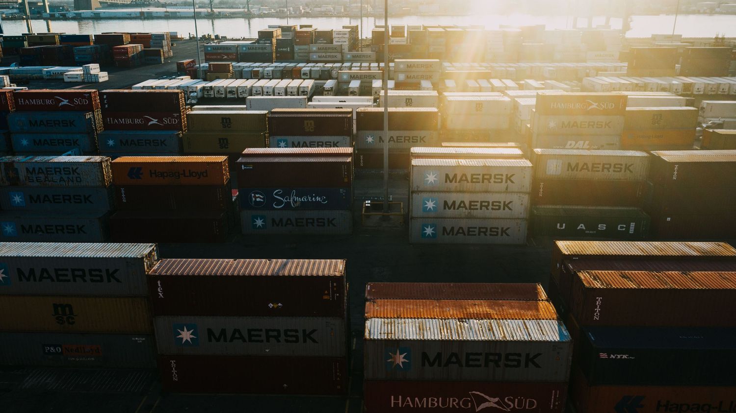 Aerial view of colorful Maersk and Hamburg Sud shipping containers stacked at a busy port terminal.