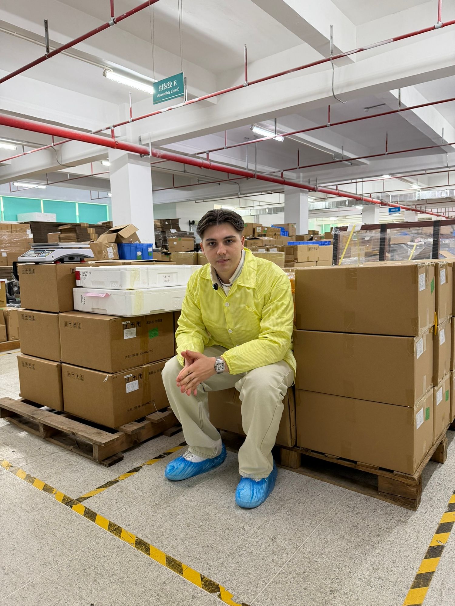 Man in yellow lab coat sitting on cardboard boxes in a warehouse facility.