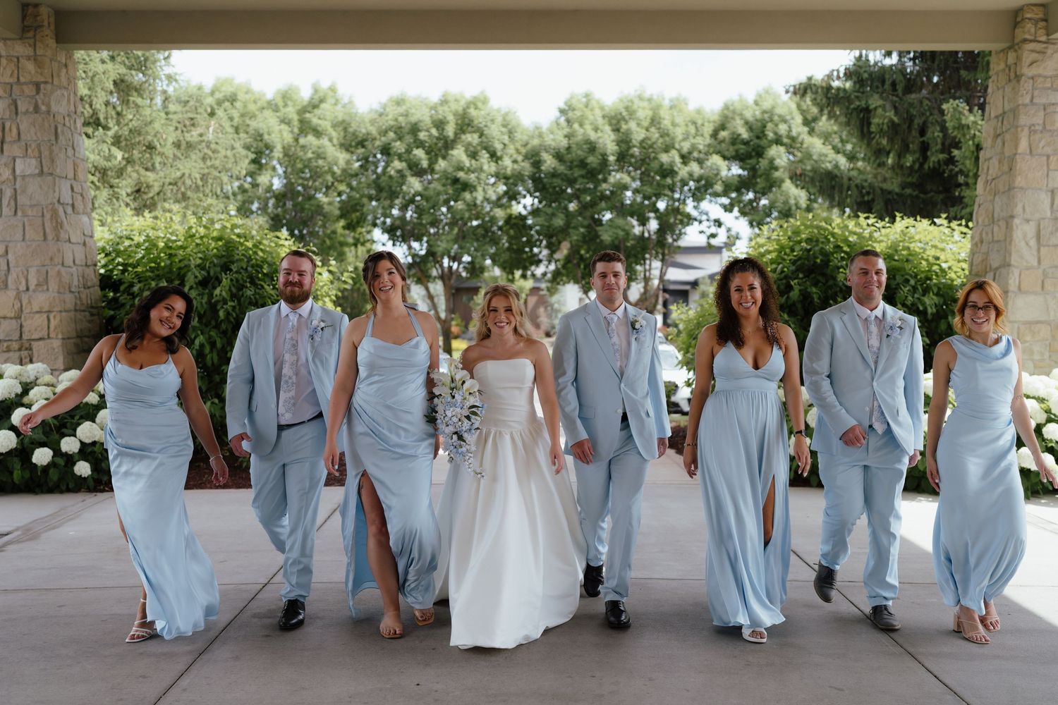 Wedding party walks together, bride in white gown, groomsmen in light blue suits, bridesmaids in matching blue dresses.