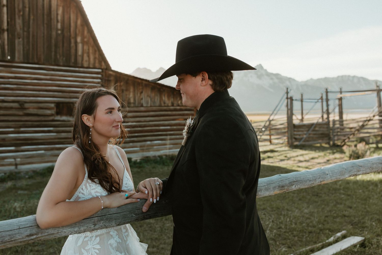 Bride and groom in cowboy attire share a romantic moment by rustic wooden fence at ranch.