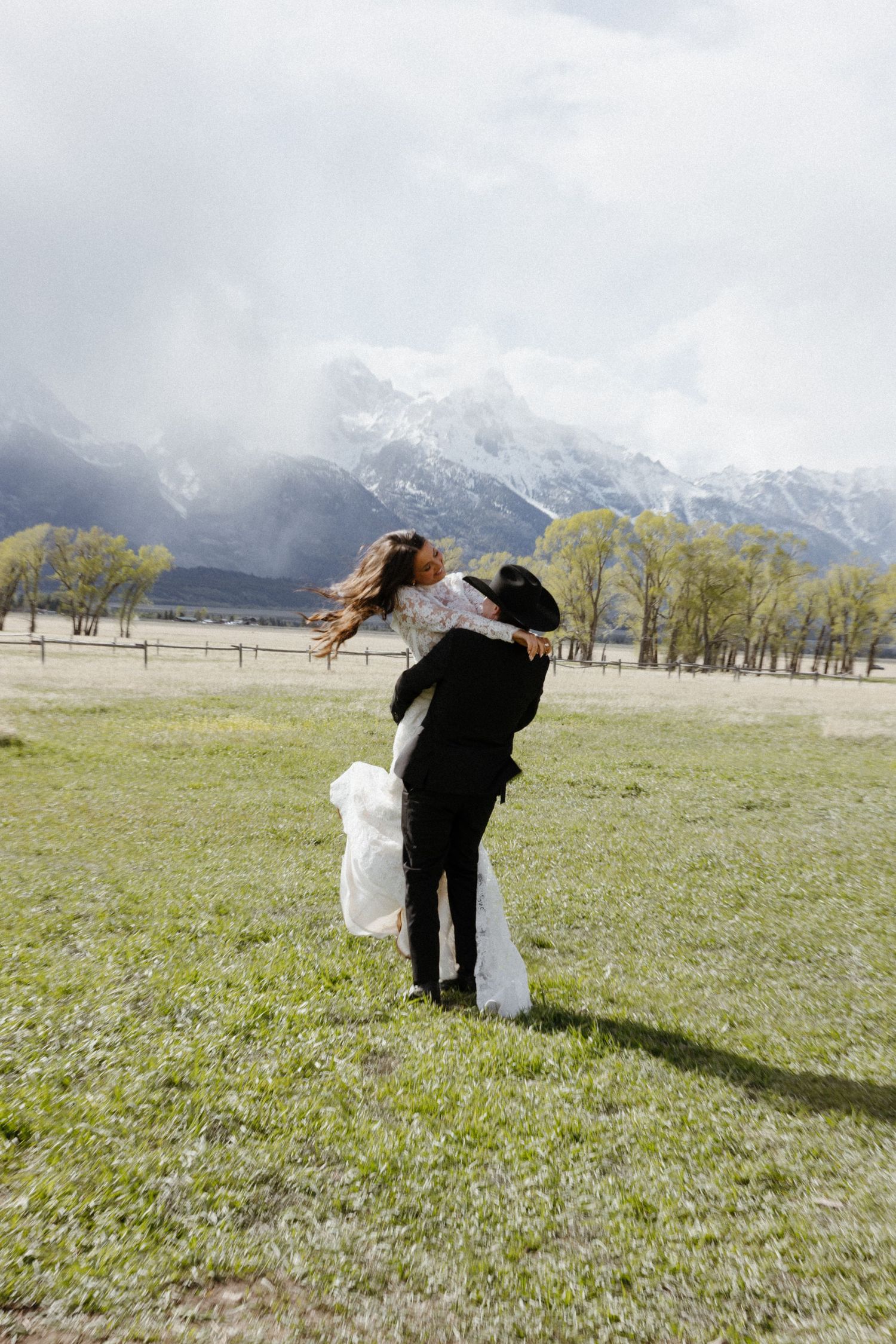 Groom lifting bride in lace wedding dress in a green meadow with snow-capped mountains behind them.