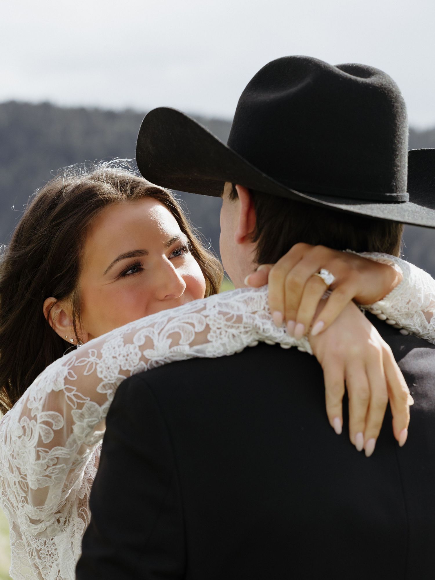 Close-up of bride with diamond ring embracing groom in black cowboy hat at outdoor western wedding.