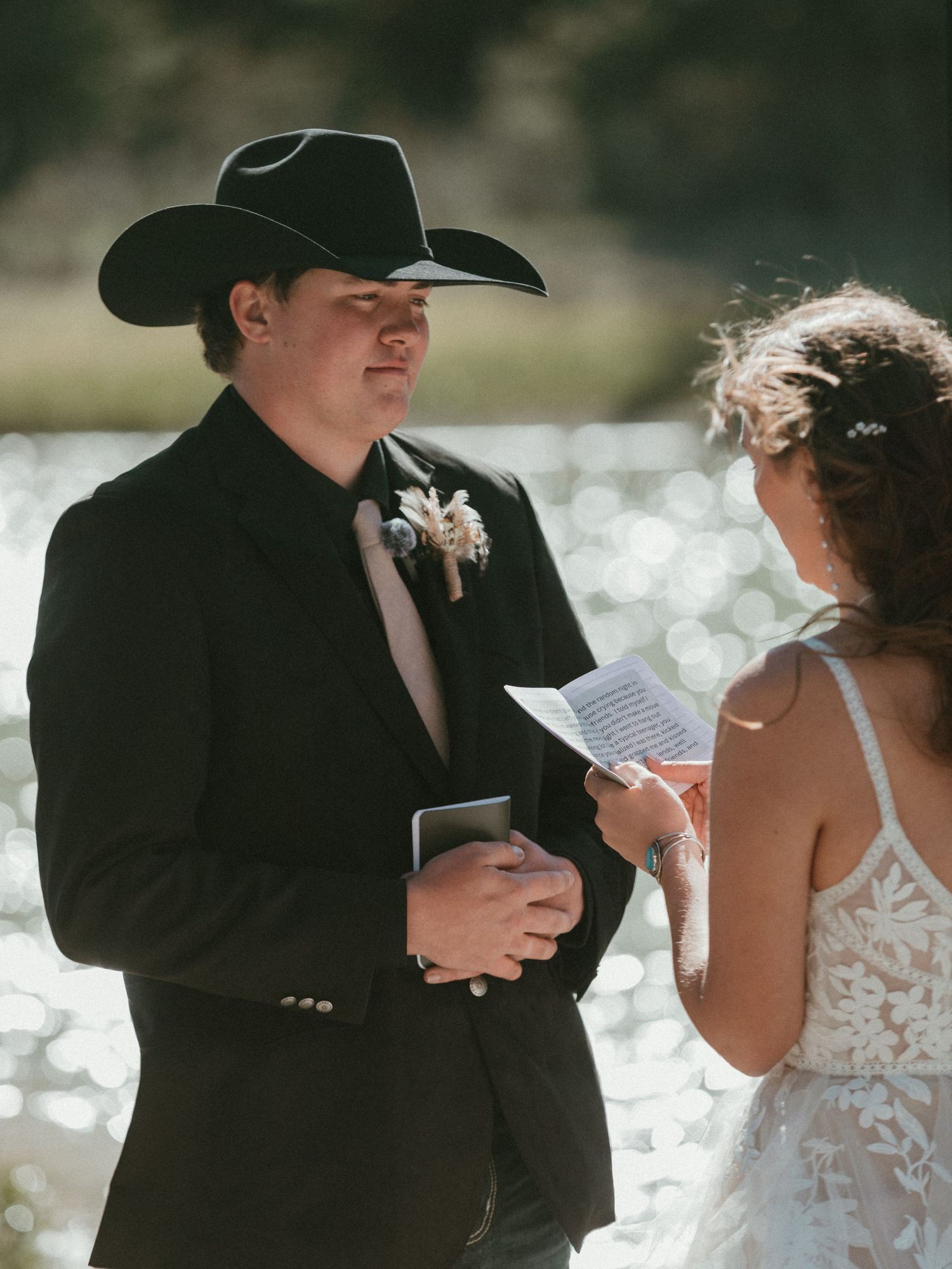 Groom in black cowboy hat and suit listens as bride reads wedding vows by a shimmering lake.